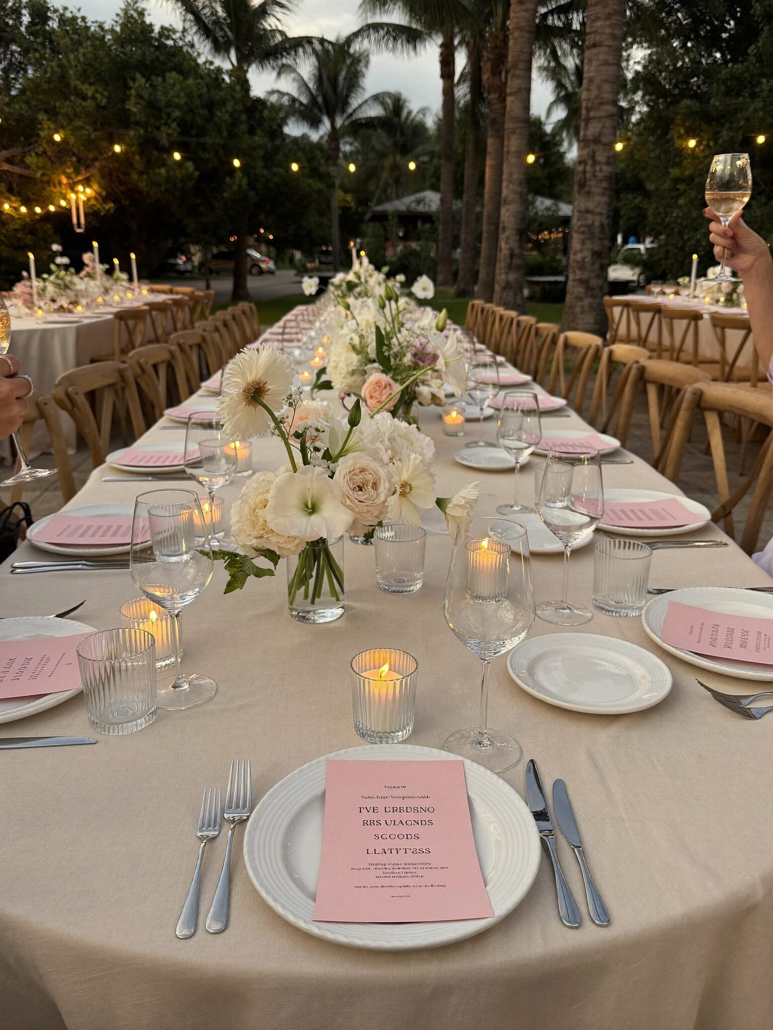 A long candlelit dinner table with ivory linens and blush flowers set outdoors under string lights