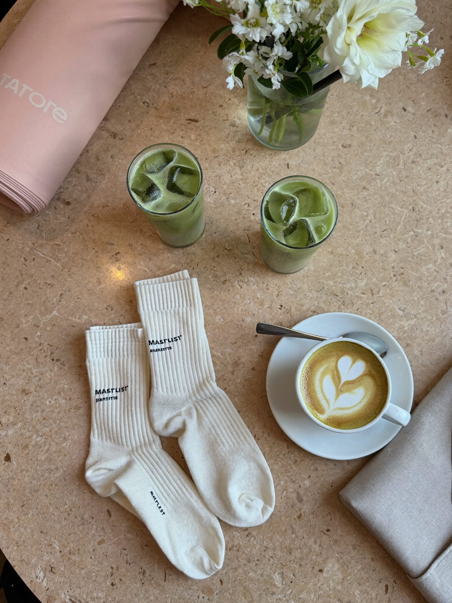 Iced matcha lattes, a cappuccino, white flowers and a rolled blush mat on a travertine table
